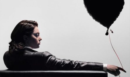 Rebecca está sentada em uma poltrona meio de costas, meio de lado. Ela usa uma jaqueta e segura um balão de gás hélio em formato de coração que flutua ao seu lado. Ela está séria e tem uma expressão sombria.